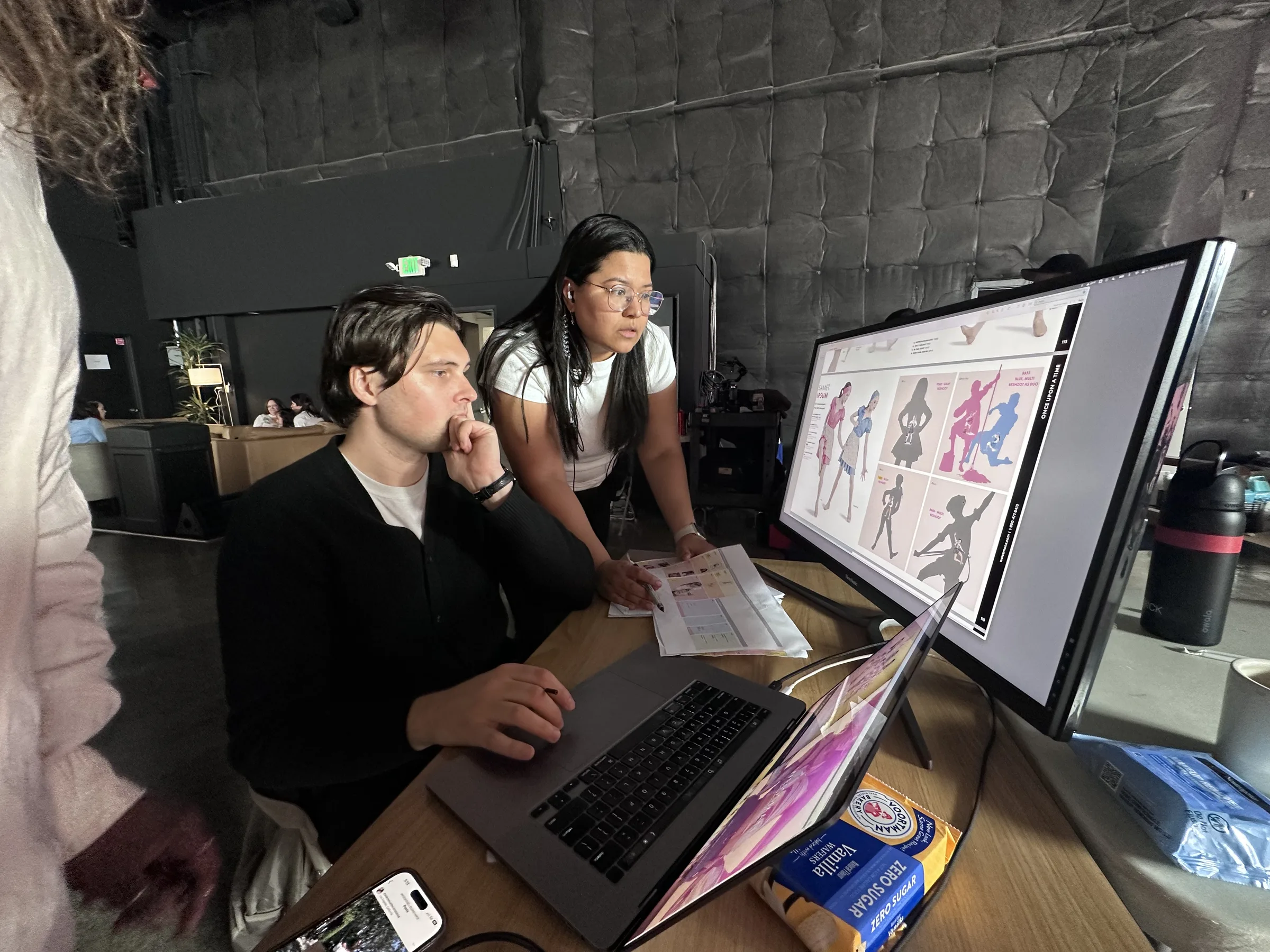 Production artist studying the monitor, chin on hand, thinking — catalog with silhouette placeholders visible on screen