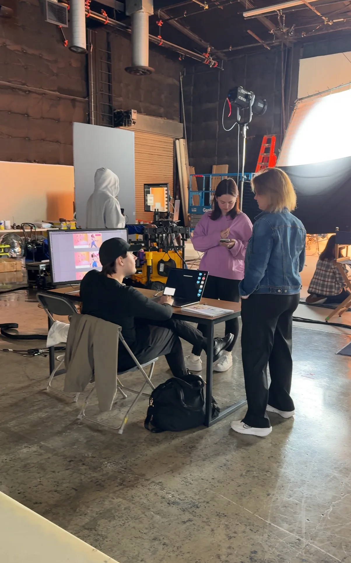 Production artist at workstation with two colleagues reviewing catalog layouts, monitors showing costume designs, full studio visible